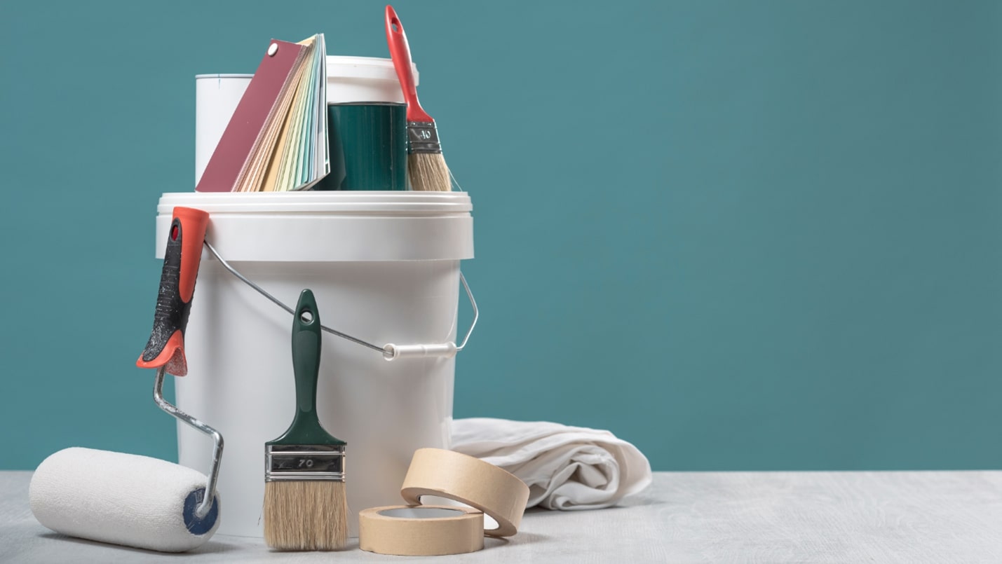 A bucket and various painting supplies, including brushes and tape, sit in front of a blue wall.